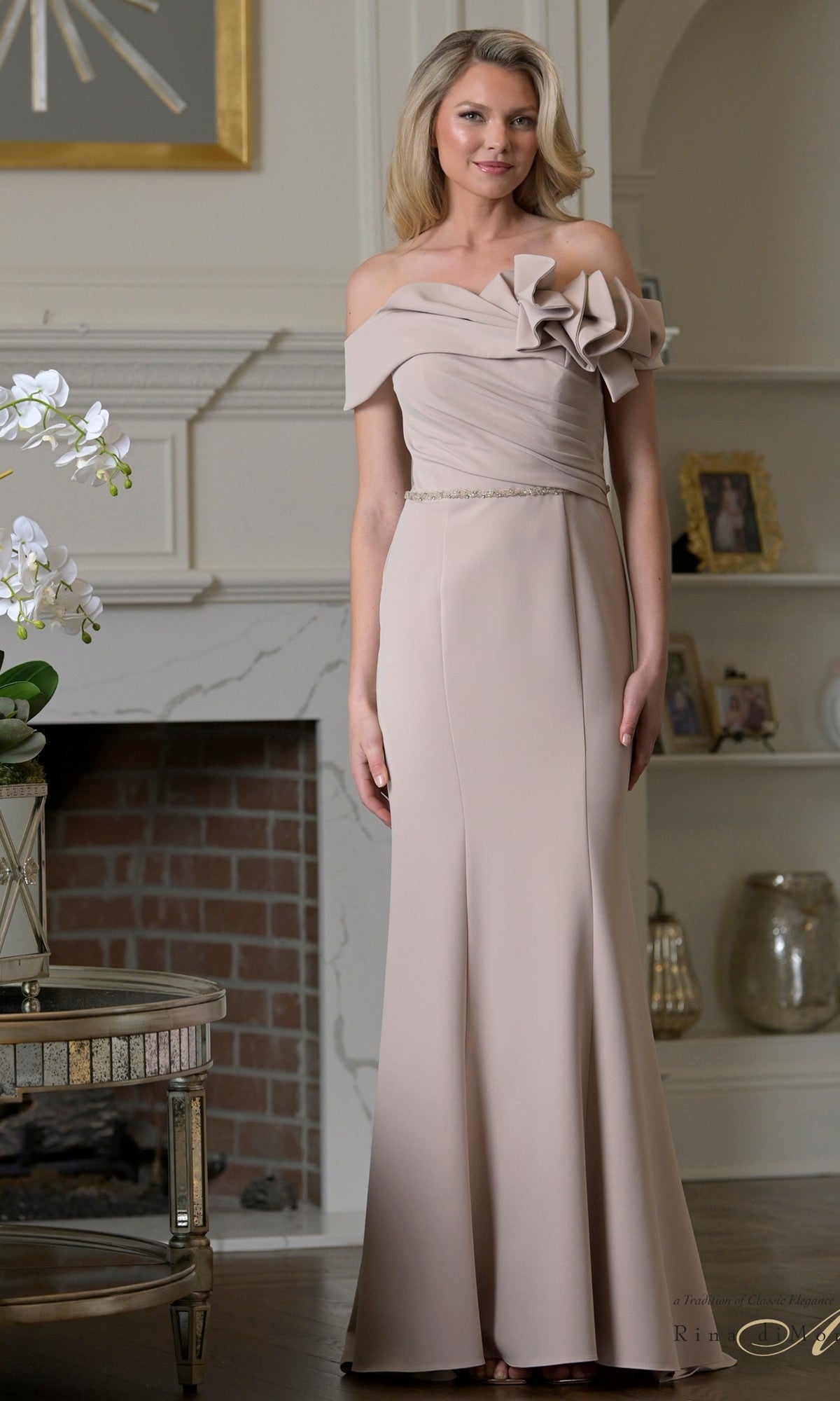 A woman wearing the Rina Di Montella RD3507 long formal dress in Champagne Gd stands in a tastefully decorated room with a fireplace and shelves in the background.